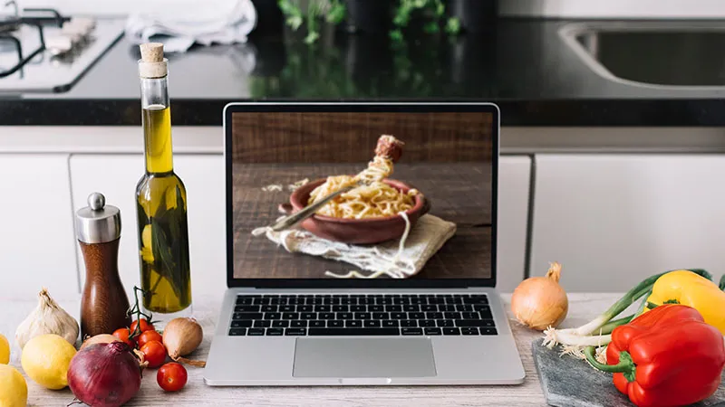 Ordenador portátil mostrando una imagen de comida casera, rodeado de ingredientes de cocina, representando estrategias de marketing digital.