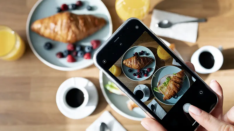 Persona tomando una foto con su móvil a un desayuno con cruasán y café, creando material visual para el plan de contenidos de un restaurante.