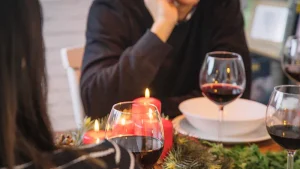 Mesa de restaurante decorada para Navidad con velas rojas, ramas de pino y copas de vino tinto, con comensales conversando de fondo.